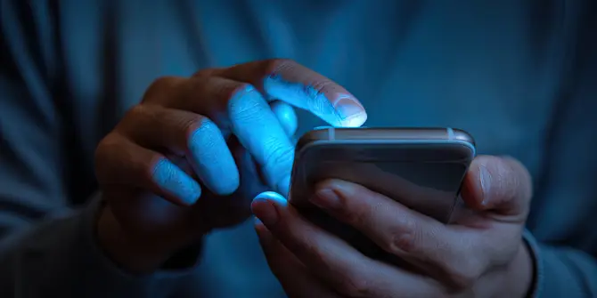 Close-Up of Person's Hands Holding Smartphone, Illuminated Screen Casting Soft Glow on Fingers, Dark Background with Blue Hue of Phone's Light, Person Interacting with Phone by Typing or Swiping